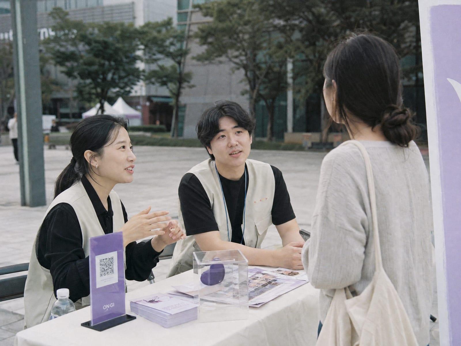 거리 캠페인 부스에서 QR 스탠드를 두고 시민과 이야기하는 비영리기관 봉사자들